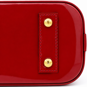 Close-up of a glossy red leather bag corner showing a stitched leather tab with two gold-tone metal studs on a white background.