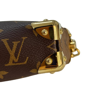 Close-up of a Louis Vuitton monogram handbag in brown coated canvas with gold-tone hardware and studs, showing the LV logo and metal corner protectors against a plain light background.