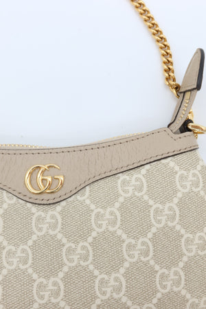 Close-up of a Gucci handbag in beige GG Supreme canvas with a beige leather trim, gold-tone interlocking G logo, and a gold chain strap on a light background.