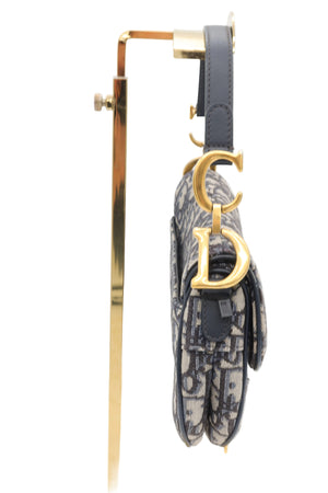 A Dior Oblique monogram shoulder bag with gold-tone hardware, hanging from a gold display stand against a white background.