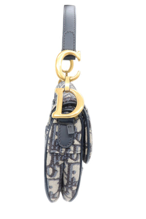 A close-up side view of a Dior saddle bag in Dior Oblique canvas, featuring a dark gray leather strap with a gold-tone 'CD' charm, held by a white-gloved hand against a white background.