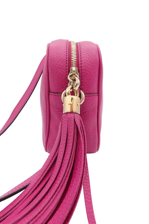 A close-up of a bright pink pebbled leather crossbody bag with a gold zipper, gold padlock charm, and a long matching leather tassel strap, shown on a white background.
