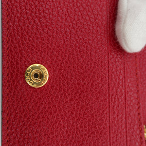 Close-up of a red textured leather handbag showing a gold-tone snap button closure and a small gold logo accent, with a white-gloved finger touching the surface.