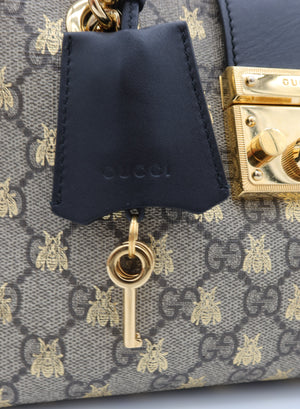 Close-up of a Gucci handbag featuring the brand's signature GG Supreme canvas with gold bee motifs, a dark navy leather hangtag, and a gold key charm.