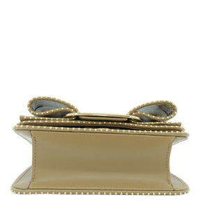 A structured beige leather clutch bag with a prominent metallic gold bow on top, decorated with gold studs along the edges, shown against a plain white background.