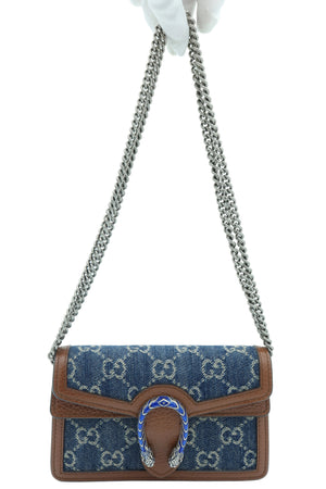 A Gucci GG Supreme canvas shoulder bag in blue and brown, held by a white-gloved hand against a light gray background. The bag features a brown leather front flap, a silver-tone chain strap, and a silver horsebit clasp embellished with blue enamel.