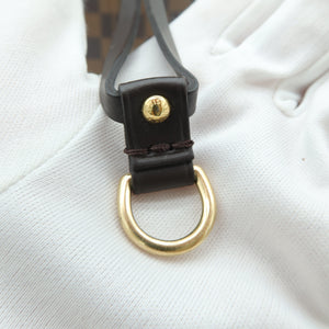 Close-up of a dark brown leather strap with a gold-tone D-ring and a small gold snap button, resting on a white textured surface with a brown checkered pattern visible in the background.