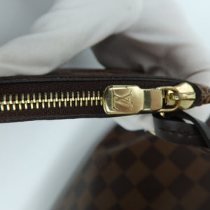 Close-up of a Louis Vuitton Damier canvas bag with a brown and black checkered pattern, featuring a gold-tone metal zipper with a gold LV logo pull, held by a white gloved hand.