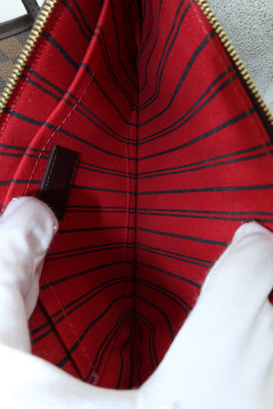 Close-up of the interior of a red fabric bag with black plaid stripes, held open by white-gloved hands. A gold zipper runs along the top edge, and a small dark brown leather brand tab is visible on the left side.