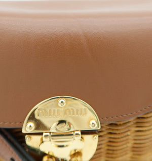Close-up of a Miu Miu shoulder bag featuring a tan leather flap with a polished gold-tone metal clasp and a woven straw base.