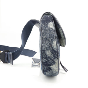 A small crossbody bag in navy blue suede with a white abstract pattern, featuring a matching navy strap and black buckle, shown from a side profile on a white background.