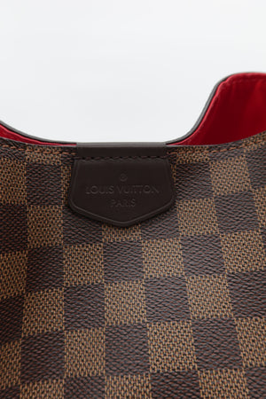 Close-up of a Louis Vuitton handbag in the Damier Ebene checkered pattern, showing a dark brown leather brand tab and a red interior lining.