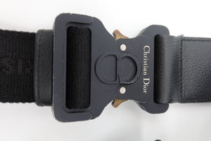 Close-up of a Christian Dior black belt buckle with a black woven strap and textured black leather section, on a white background.