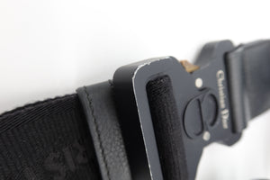 Close-up of a black tactical belt with a matte black metal buckle, featuring a textured webbing strap and a black velcro fastening system. The buckle has 'Christian Dior' printed in white on the side.