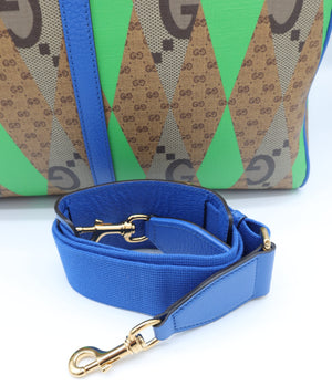 A Gucci tote bag in green and brown GG monogram canvas with a bright blue leather shoulder strap and gold-tone hardware, shown on a white background.