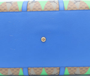 A close-up of a Gucci handbag showing the bottom flap, featuring a vibrant blue pebbled leather center with a gold-tone snap button, and green and beige GG Supreme patterned fabric borders on the top and bottom.