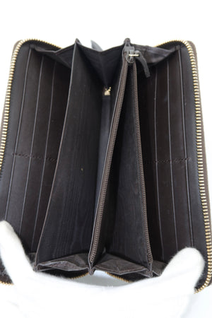 An open dark brown monogram wallet or clutch shown from the inside, with multiple card slots, a central zip compartment, and gold-tone zippers. White-gloved hands are visible holding the item against a plain white background.