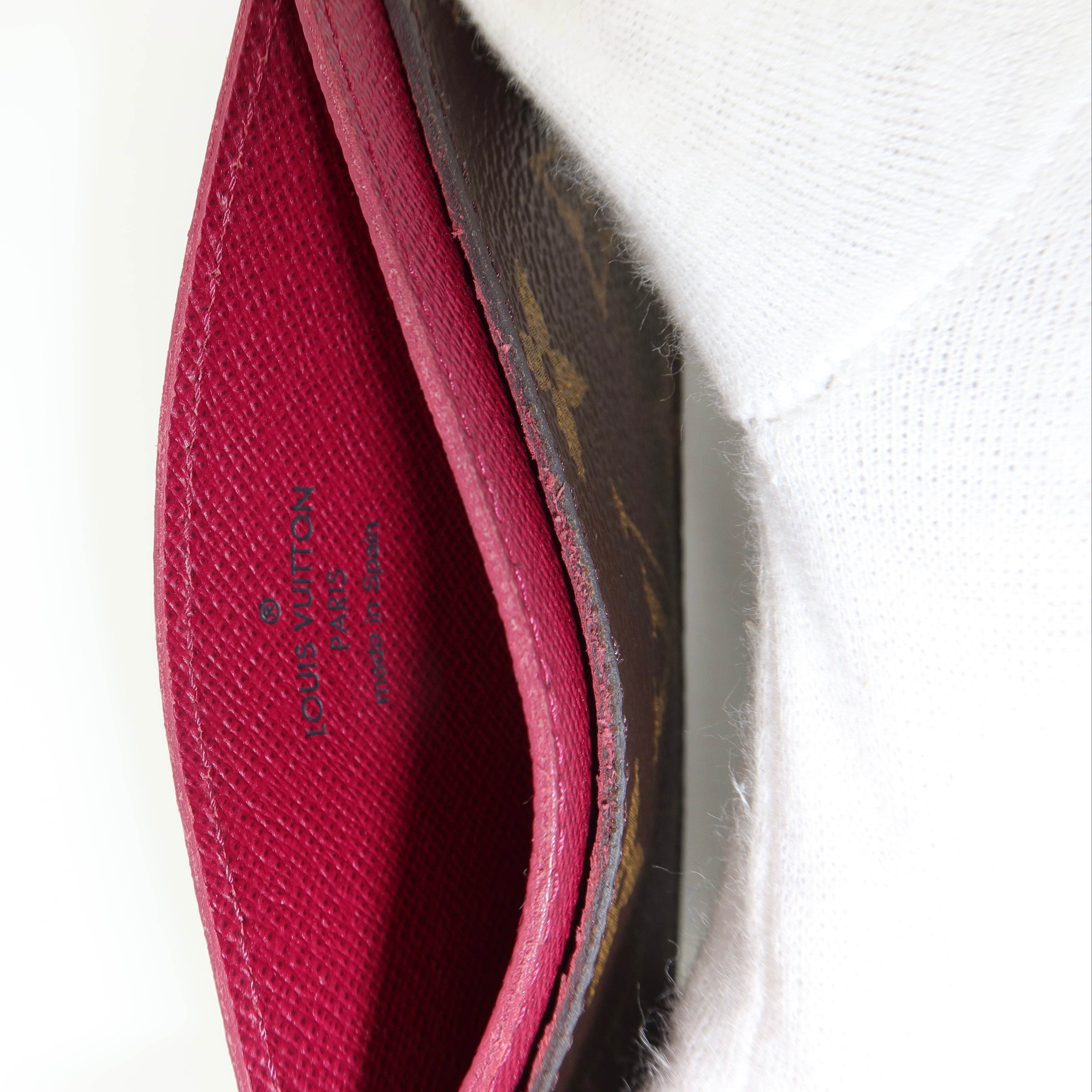 Close-up of an open Louis Vuitton wallet showing a bright magenta-pink interior lining and the brand name printed inside, next to a white textured fabric background.