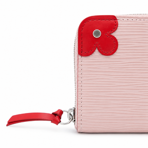 A close-up of a light pink textured leather zip-around wallet with a bright red leather flower-shaped accent and a matching red pull tab on a white background.