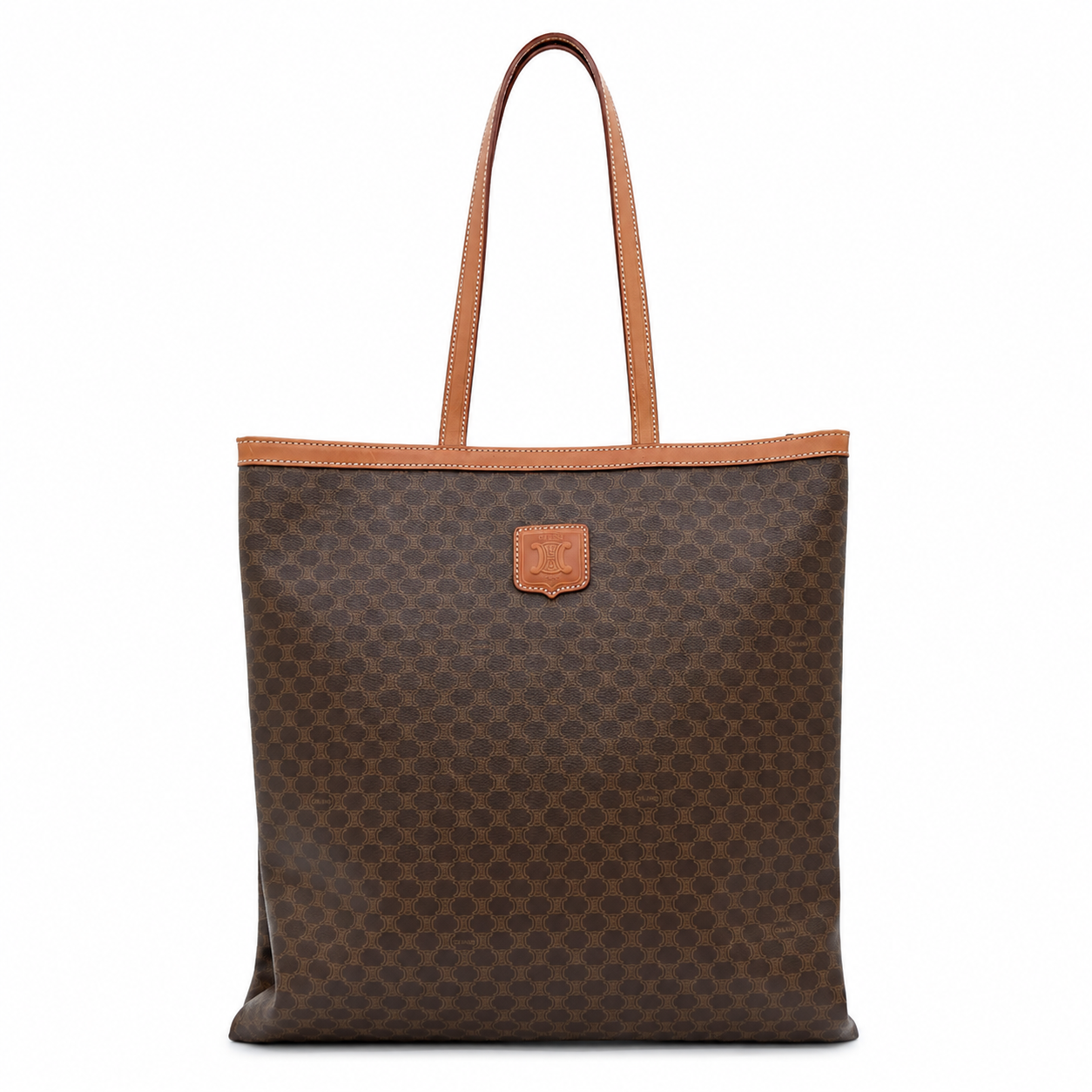 A brown monogram canvas tote bag with tan leather trim and handles, featuring a small tan leather logo patch on the front, set against a plain white background.