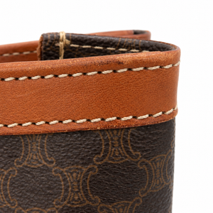 Close-up of a brown leather and dark brown monogram coated canvas bag corner with white stitching on a white background.