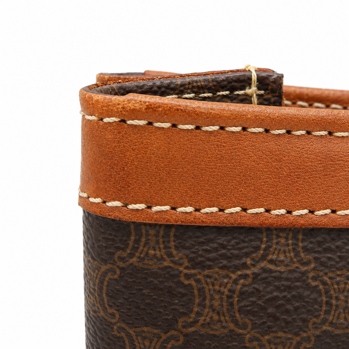 Close-up detail of a brown monogram leather accessory, showing a tan leather trim band with white stitching and a dark brown patterned body on a white background.