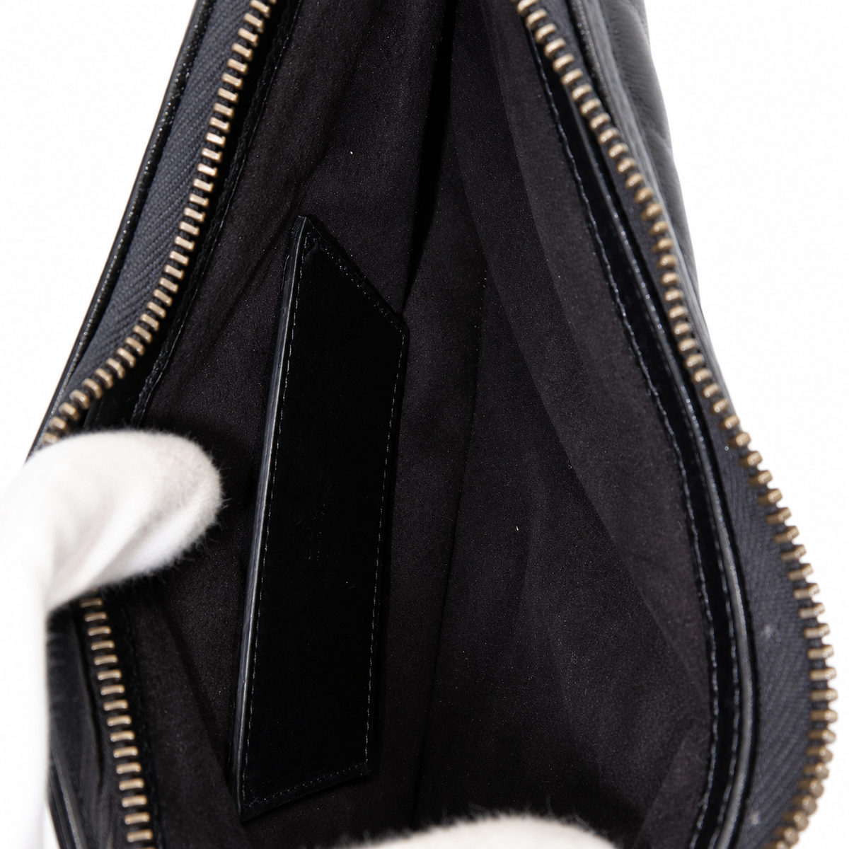 Close-up of the interior of a black leather bag with a black fabric lining, gold-toned zipper, and a white-gloved hand holding it open against a white background.