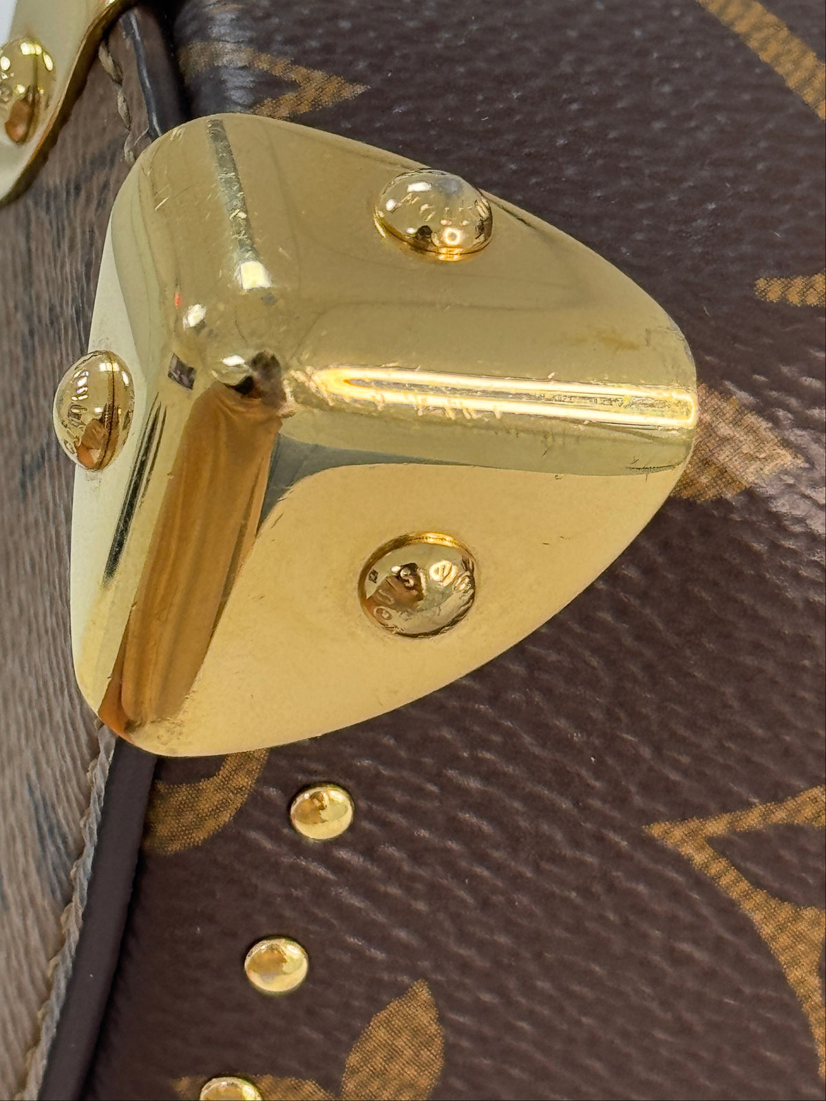 Close-up of a Louis Vuitton monogram bag corner showing the brown coated canvas with gold-tone LV monogram pattern and a polished gold metal corner protector with screws.