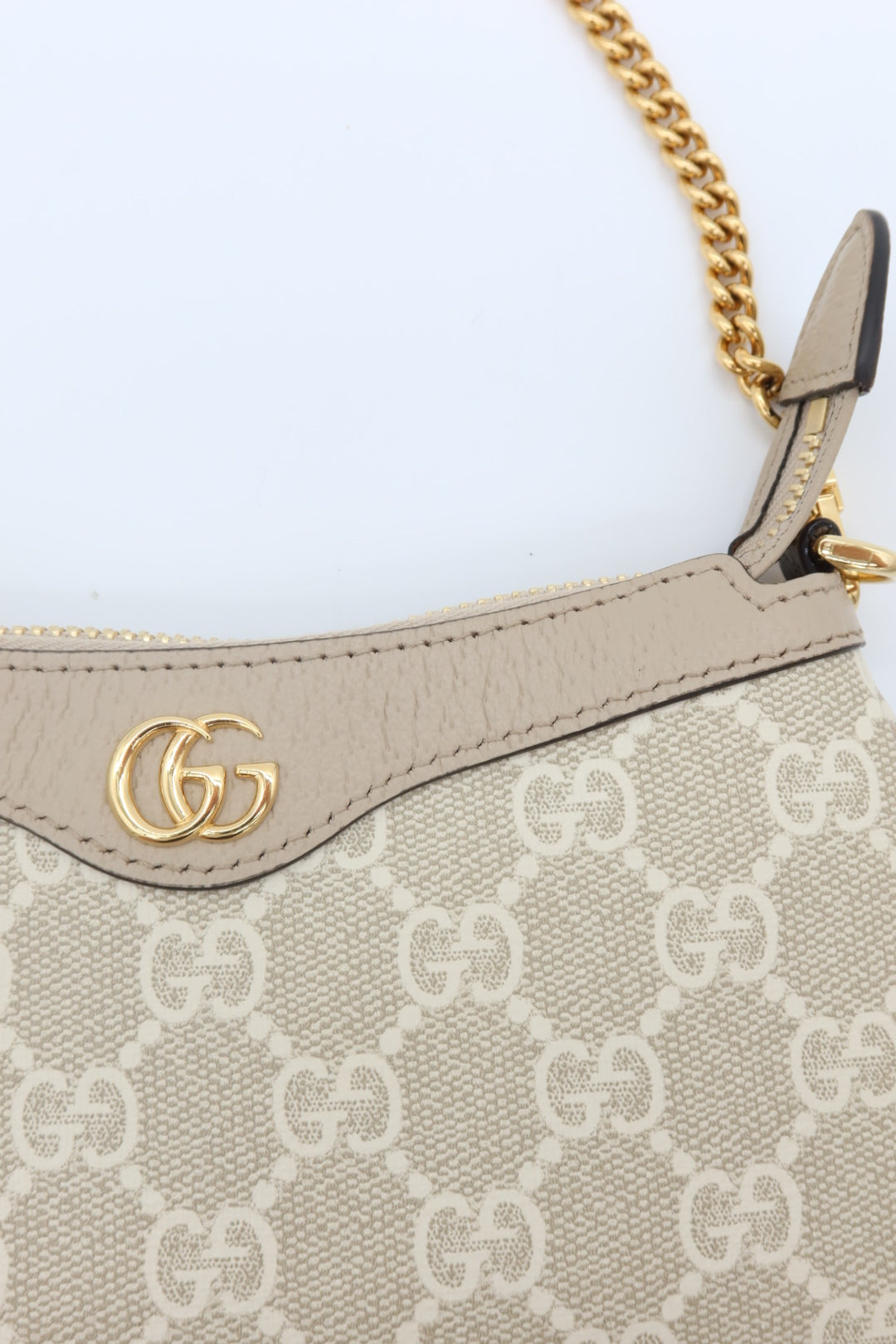 Close-up of a Gucci handbag in beige GG Supreme canvas with a beige leather trim, gold-tone interlocking G logo, and a gold chain strap on a light background.