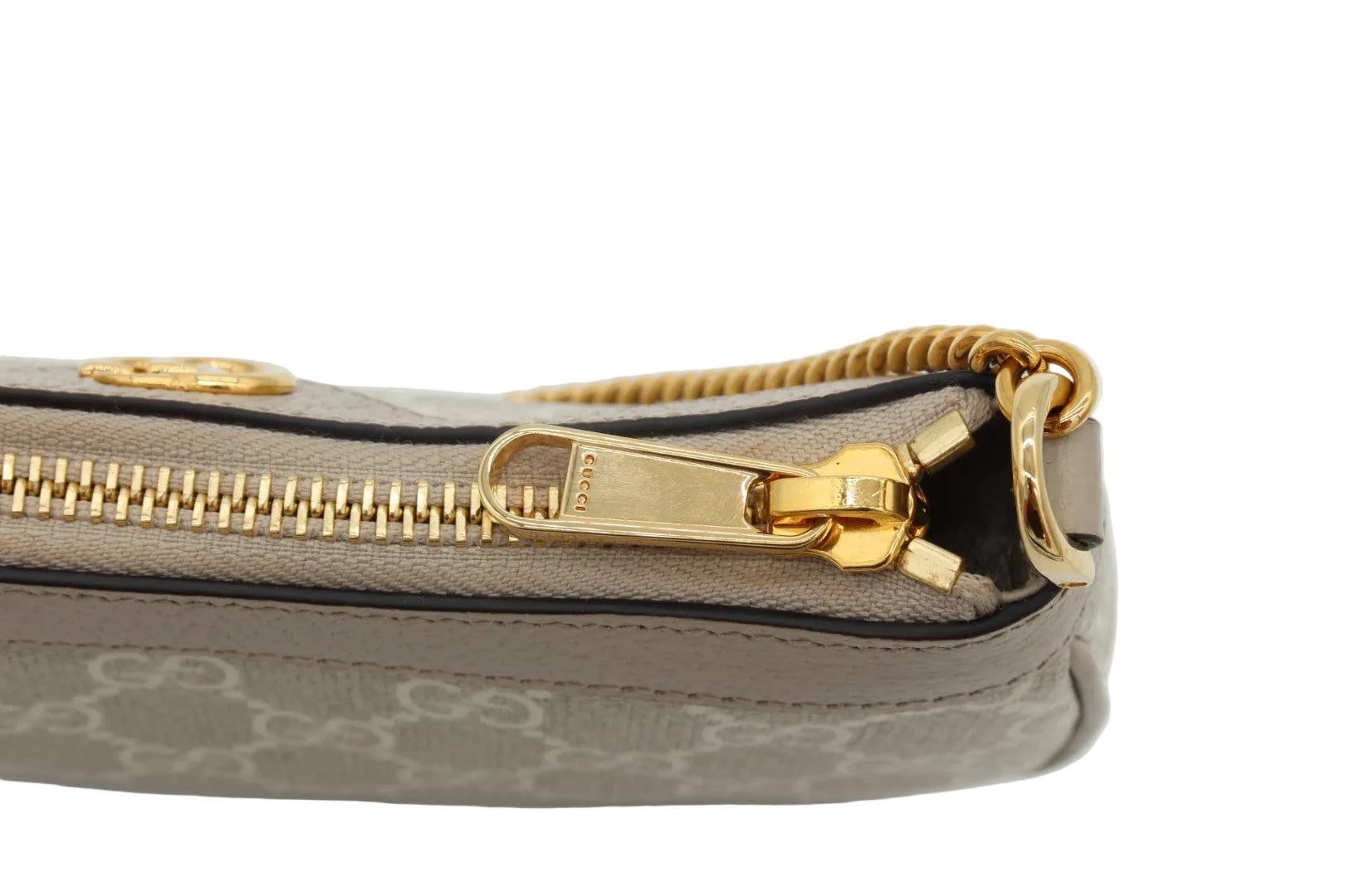 Close-up of a beige and brown monogram clutch or pouch with a gold zipper and gold hardware, featuring the Gucci logo pattern and a gold zipper pull engraved with 'GUCCI'.