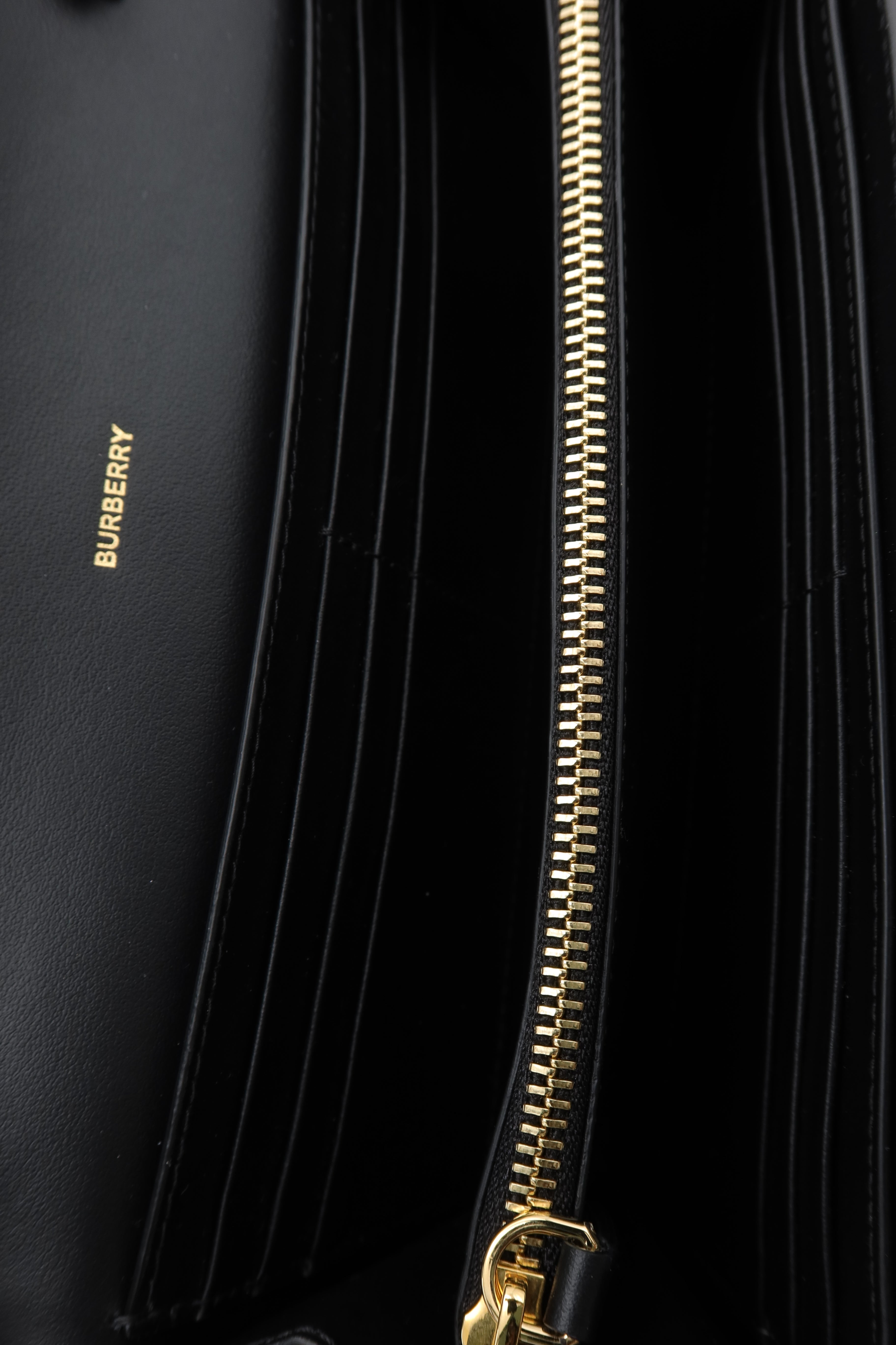 Close-up of a black leather wallet or clutch with multiple vertical card slots, a gold zipper, and a small gold BURBERRY logo on the left side.