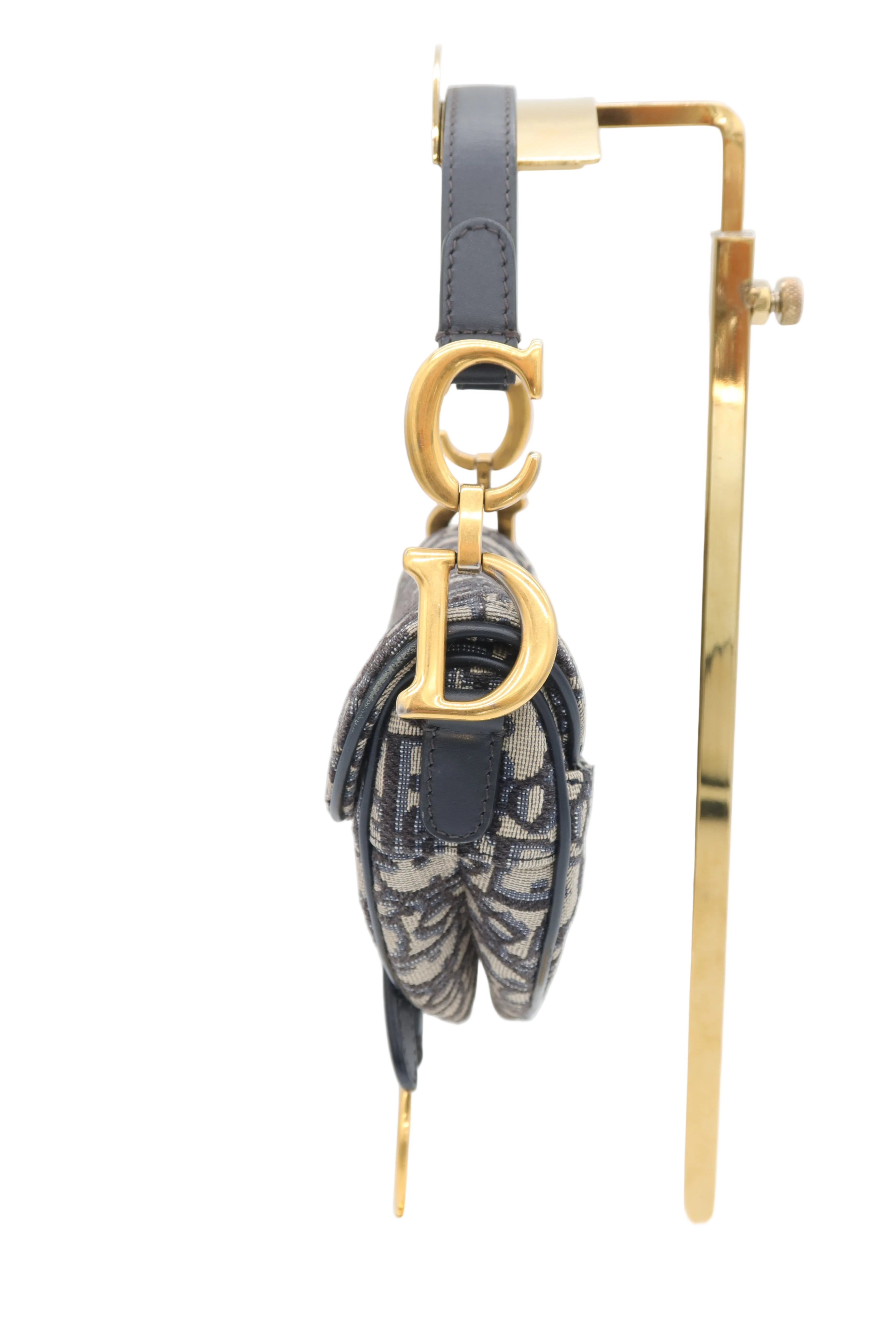 A Dior monogram shoulder bag in navy blue leather and canvas, featuring a gold-tone Dior logo charm, hanging from a gold display stand against a white background.