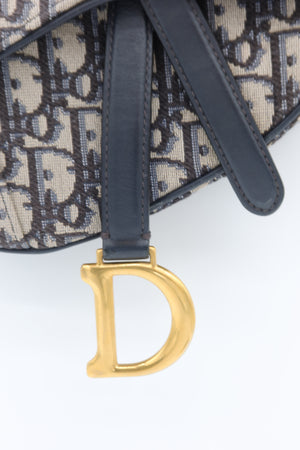 Close-up of a Dior monogram bag with dark navy leather strap and a prominent gold-tone 'D' charm on a white background.