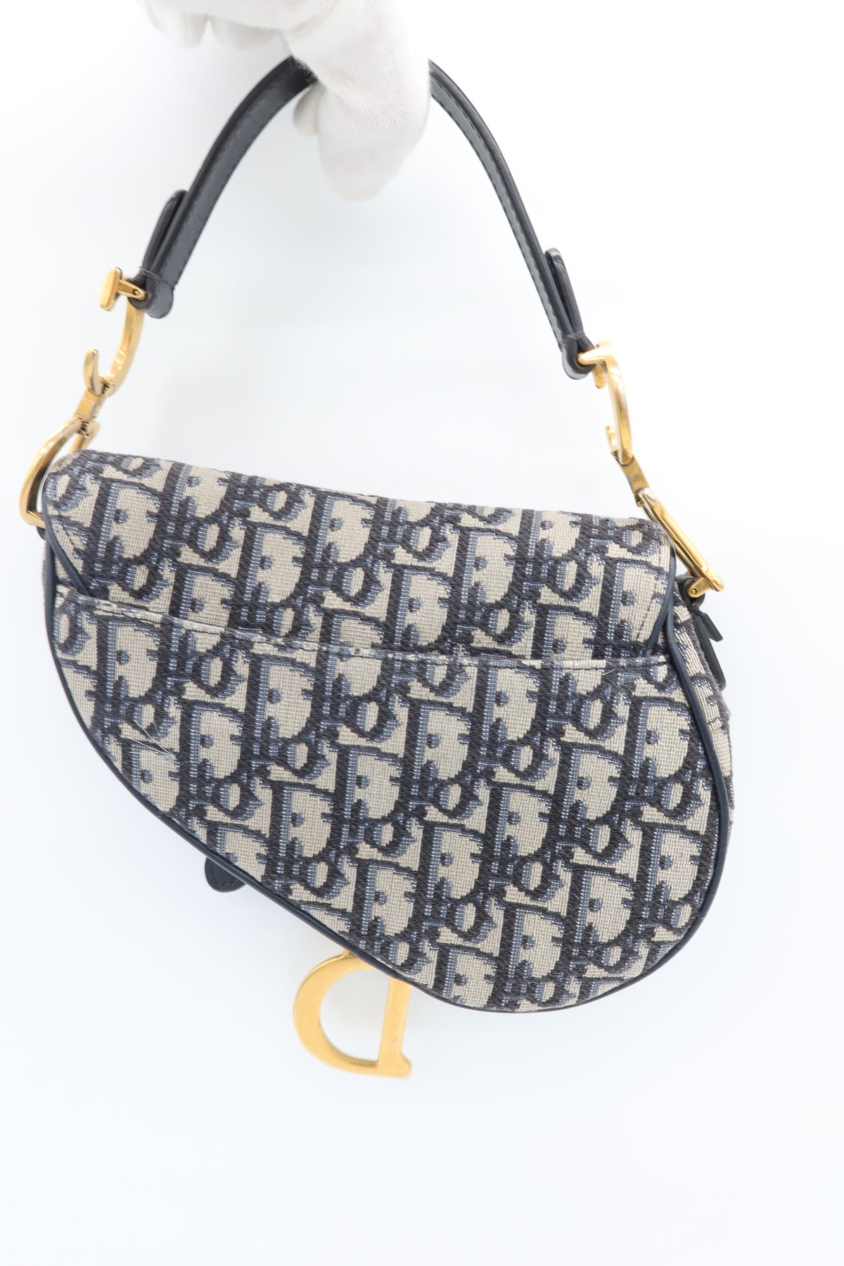A Dior Saddle bag in the signature Oblique monogram canvas, held by a white-gloved hand against a plain white background. The bag features a dark navy blue and beige monogram pattern, black leather trim and handle, and gold-tone hardware.