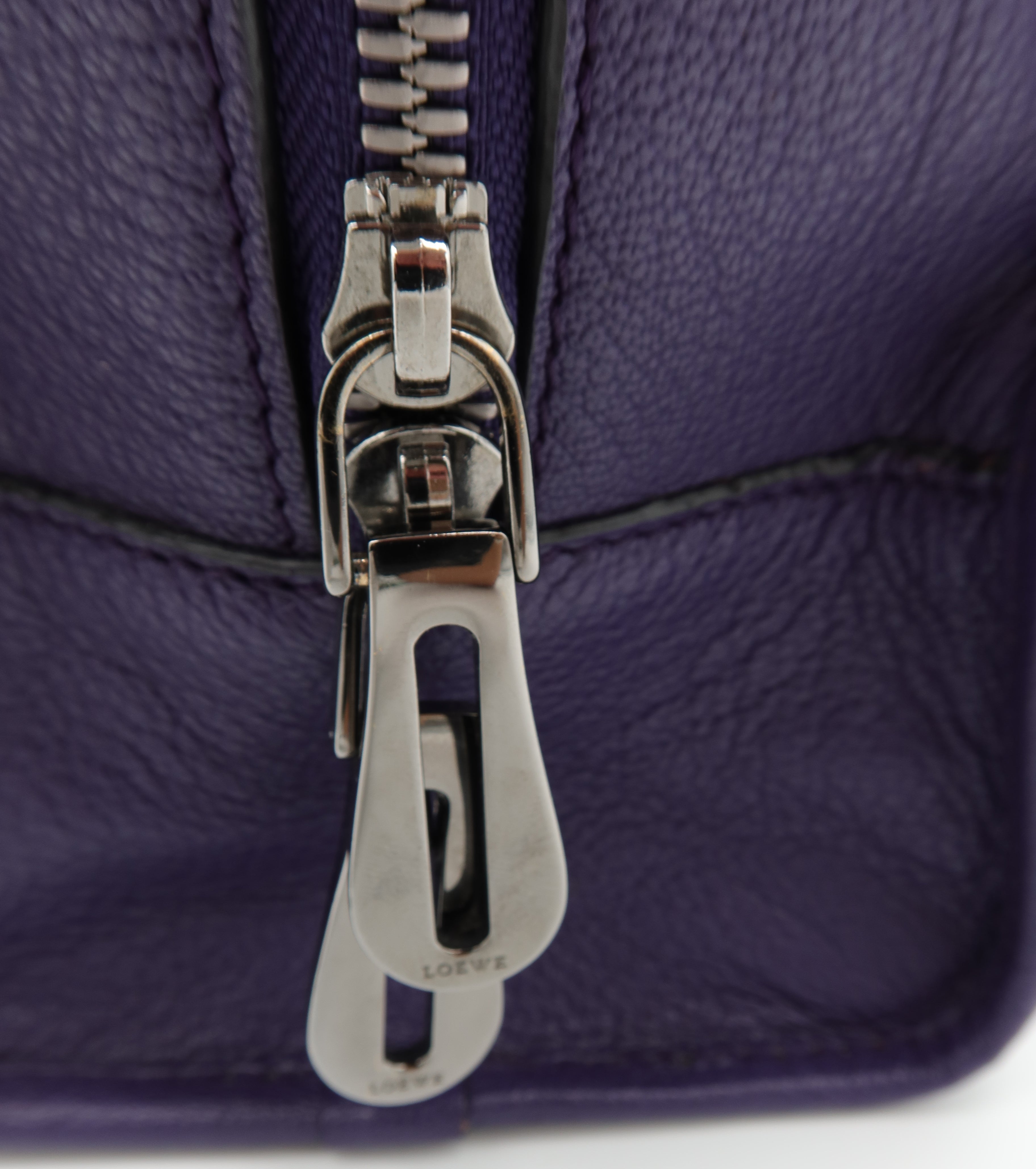 Close-up of a purple textured leather zipper pull with a silver-tone metal slider and a small embossed brand mark on the pull tab.