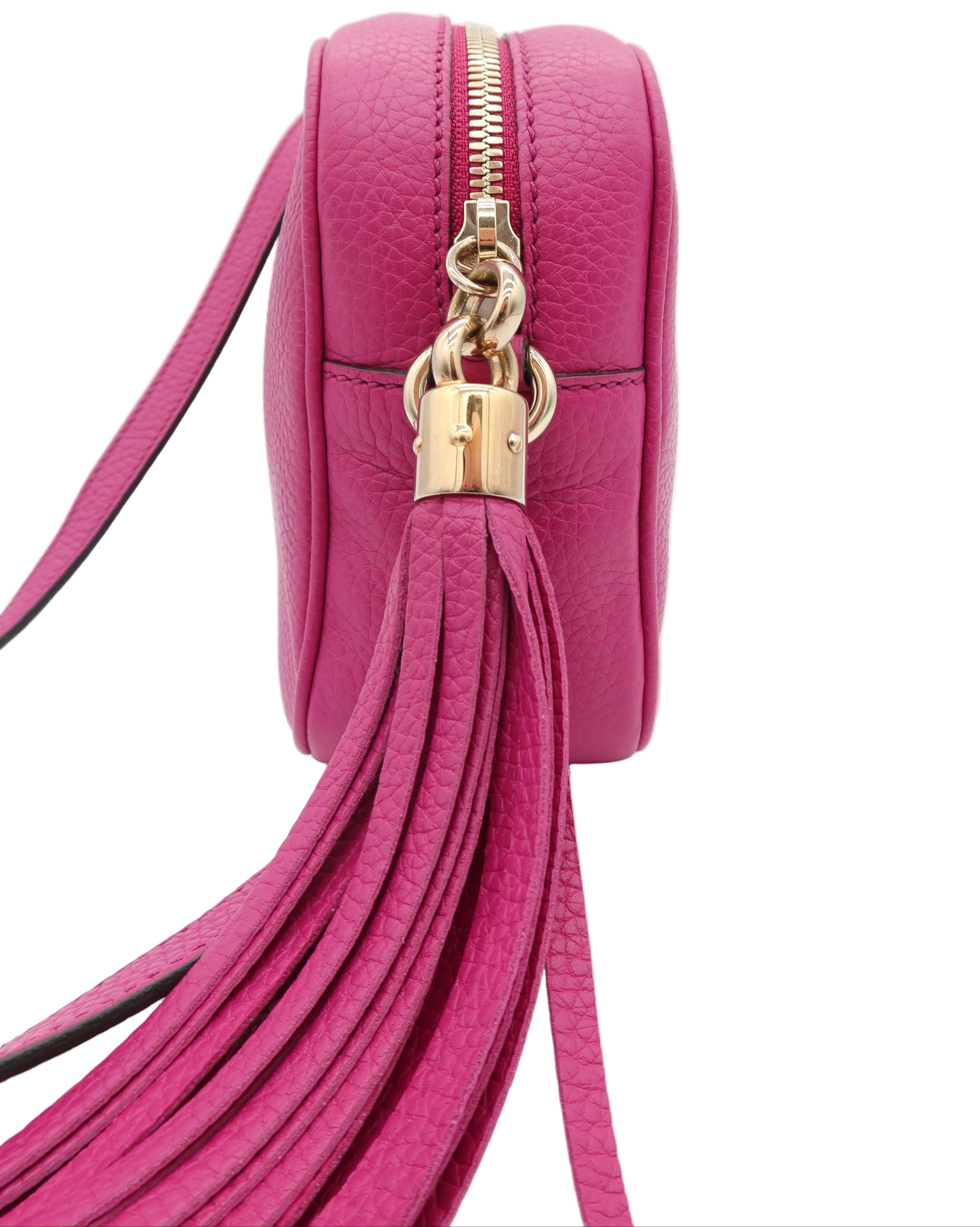 A close-up of a bright pink pebbled leather crossbody bag with a gold zipper, gold padlock charm, and a long matching leather tassel strap, shown on a white background.