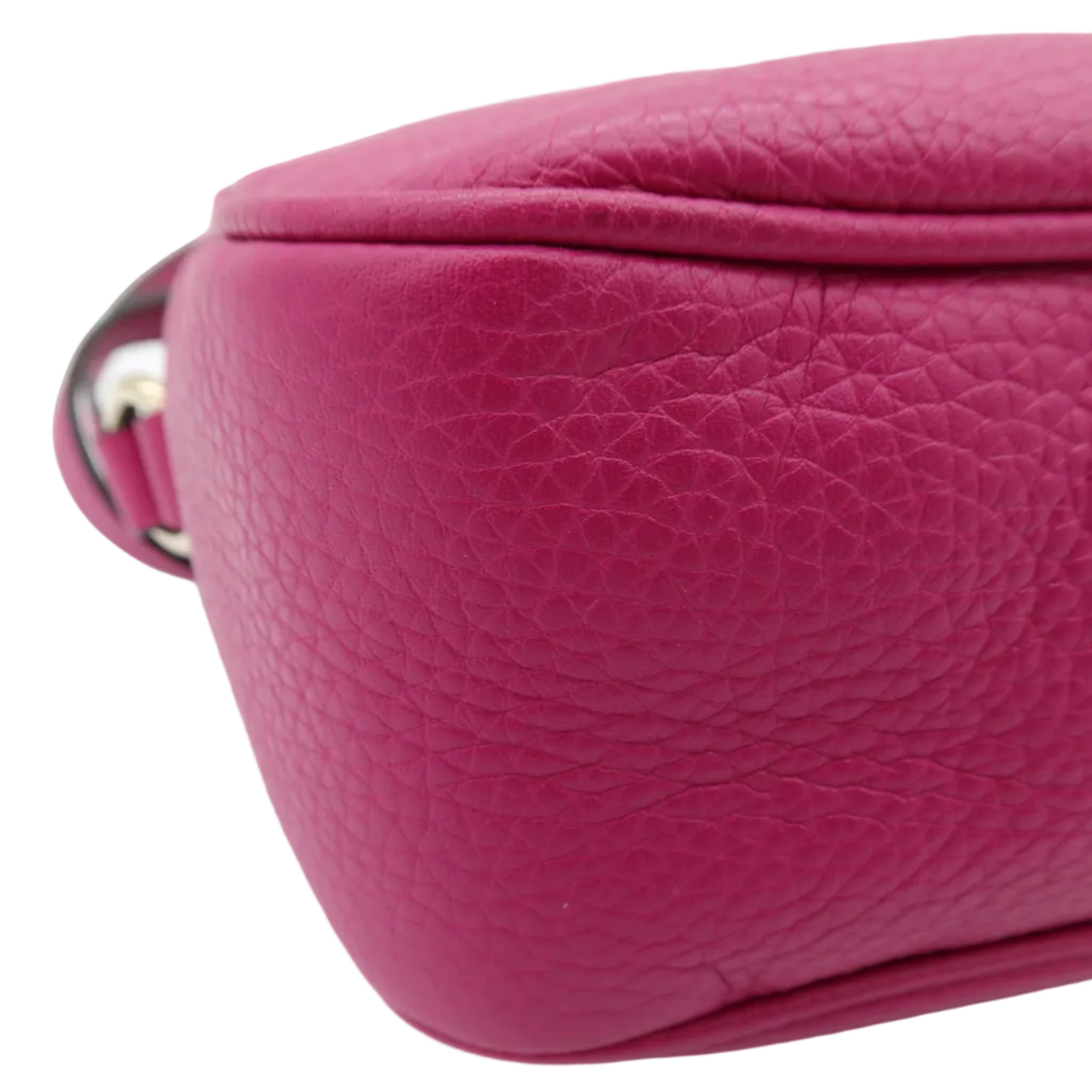 A close-up of a bright pink pebbled leather bag corner showing the texture and a small portion of the strap with a silver metal ring, on a white background.
