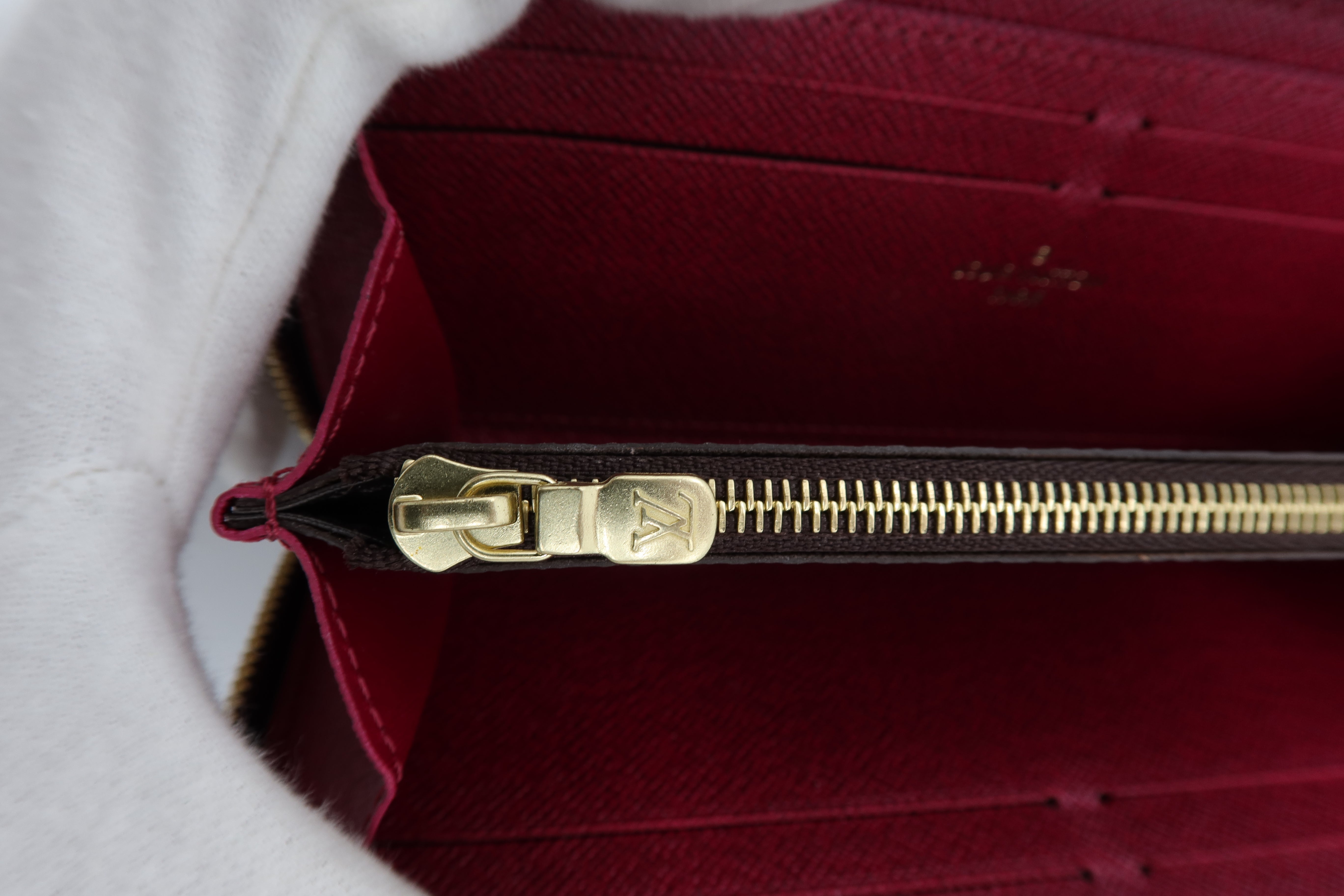 Close-up of a Louis Vuitton wallet interior showing a deep burgundy red interior, gold-tone zipper with a gold-tone metal pull tab, and a white textured fabric lining or sleeve on the left side.