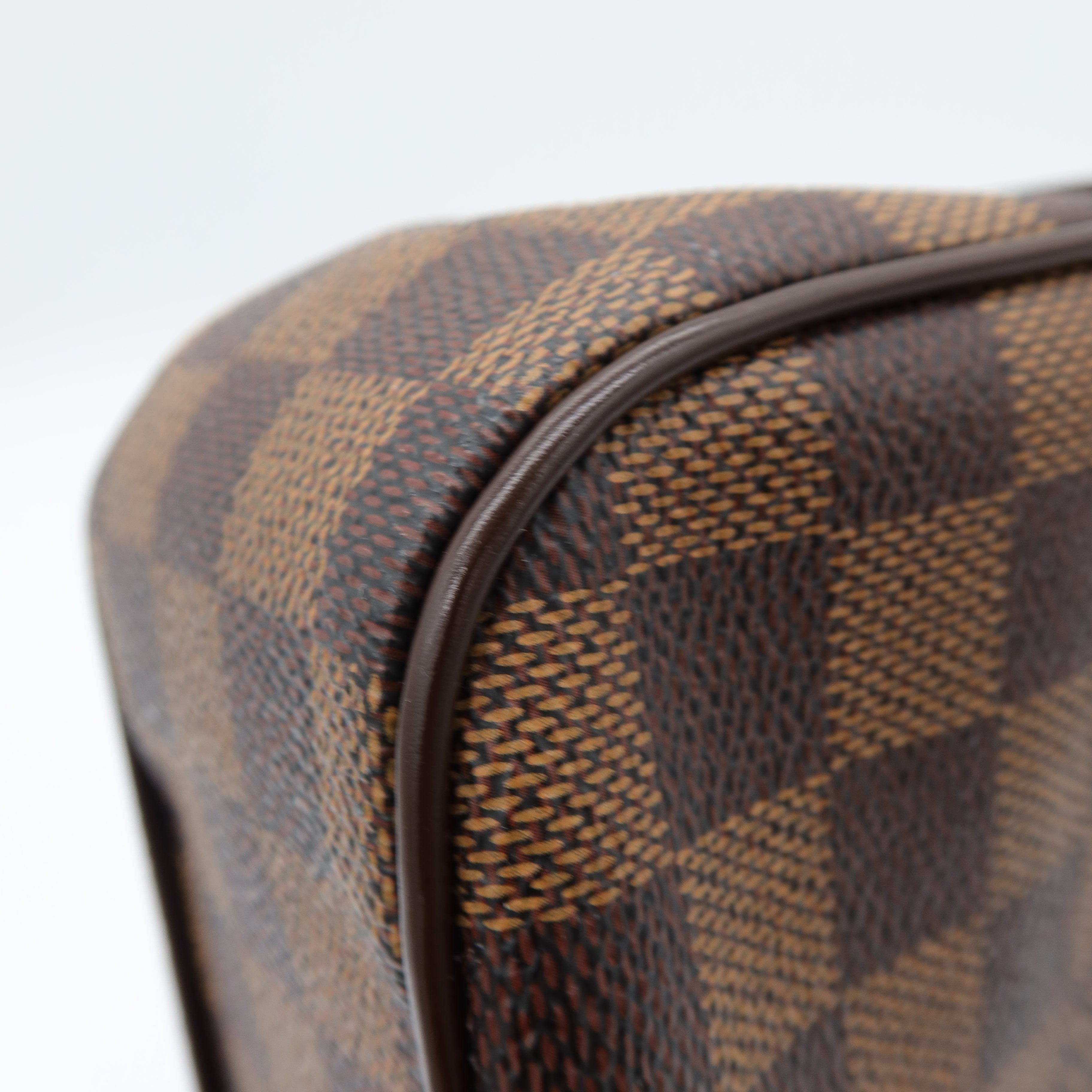 Close-up of a Louis Vuitton Damier canvas bag corner showing the brown and tan checkered pattern and dark brown leather trim against a white background.