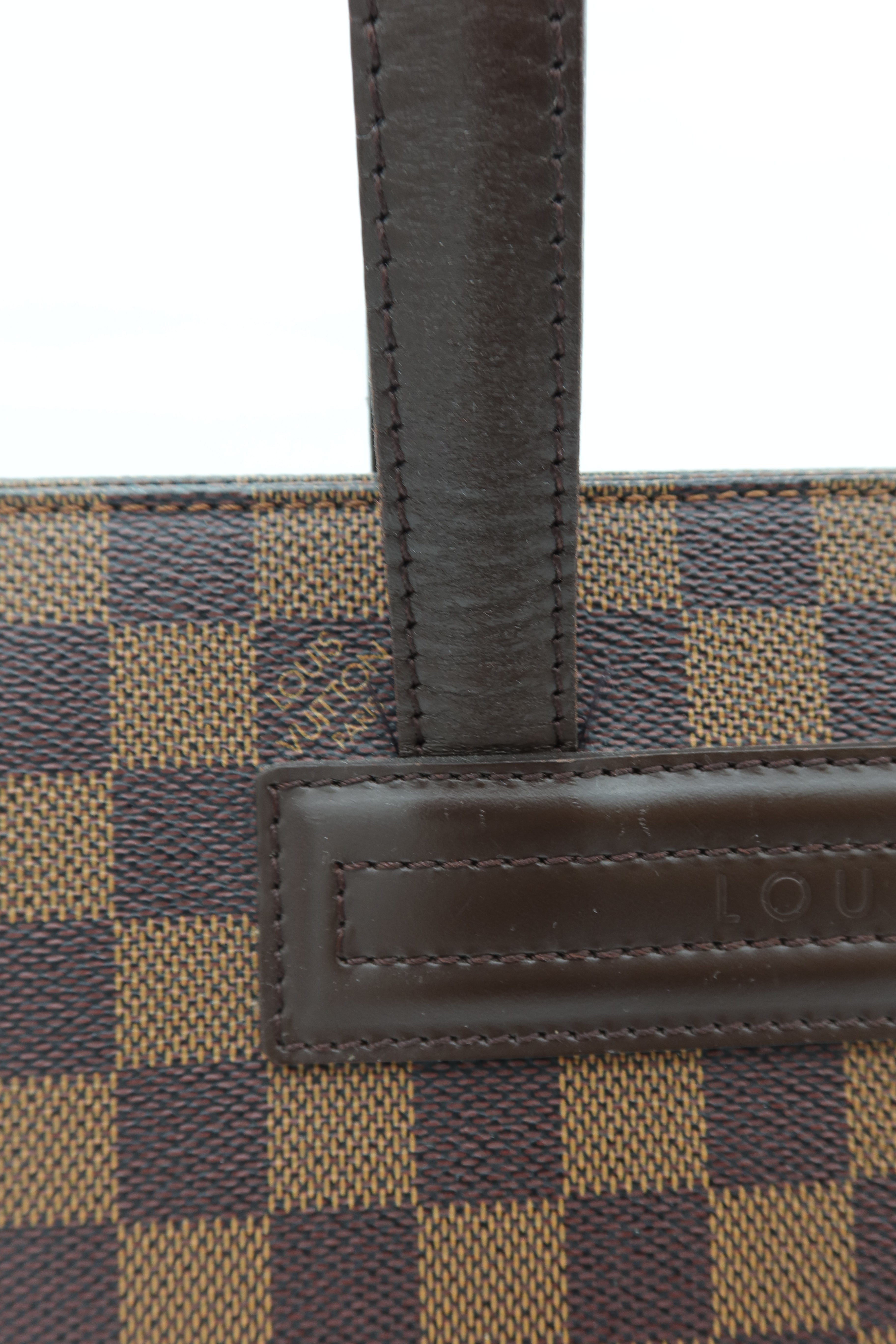 Close-up of a Louis Vuitton handbag featuring the brand's Damier check pattern in brown and beige canvas, with a dark brown leather strap and a leather patch embossed with the Louis Vuitton name.
