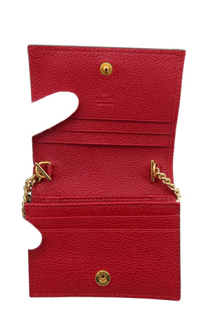 A red textured leather wallet held open by a white-gloved hand, showing gold-tone hardware, card slots, and chain details against a plain white background.