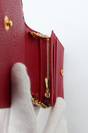A close-up of a red textured leather wallet or card holder with gold-tone hardware, held by white-gloved hands against a plain white background.