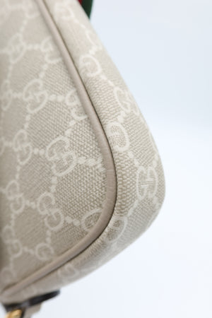 Close-up of a beige canvas shoulder bag with a repeating white monogram pattern, showing the corner and curved edge against a light background.