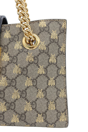 Close-up of a Gucci handbag featuring the signature GG monogram canvas with gold bee motifs and a gold chain strap ring, on a light gray background.