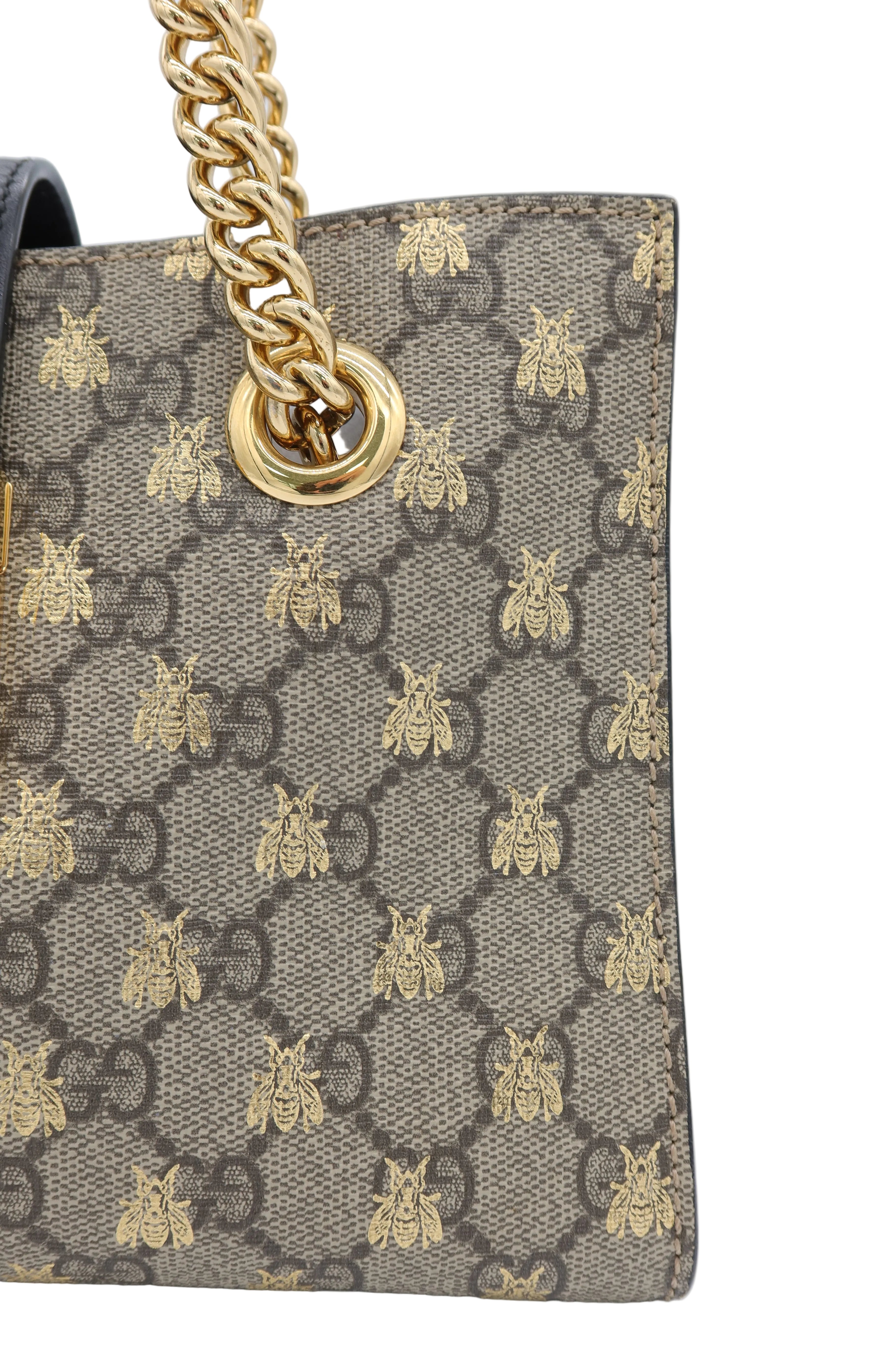 Close-up of a Gucci handbag featuring the signature GG monogram canvas with gold bee motifs and a gold chain strap ring, on a light gray background.