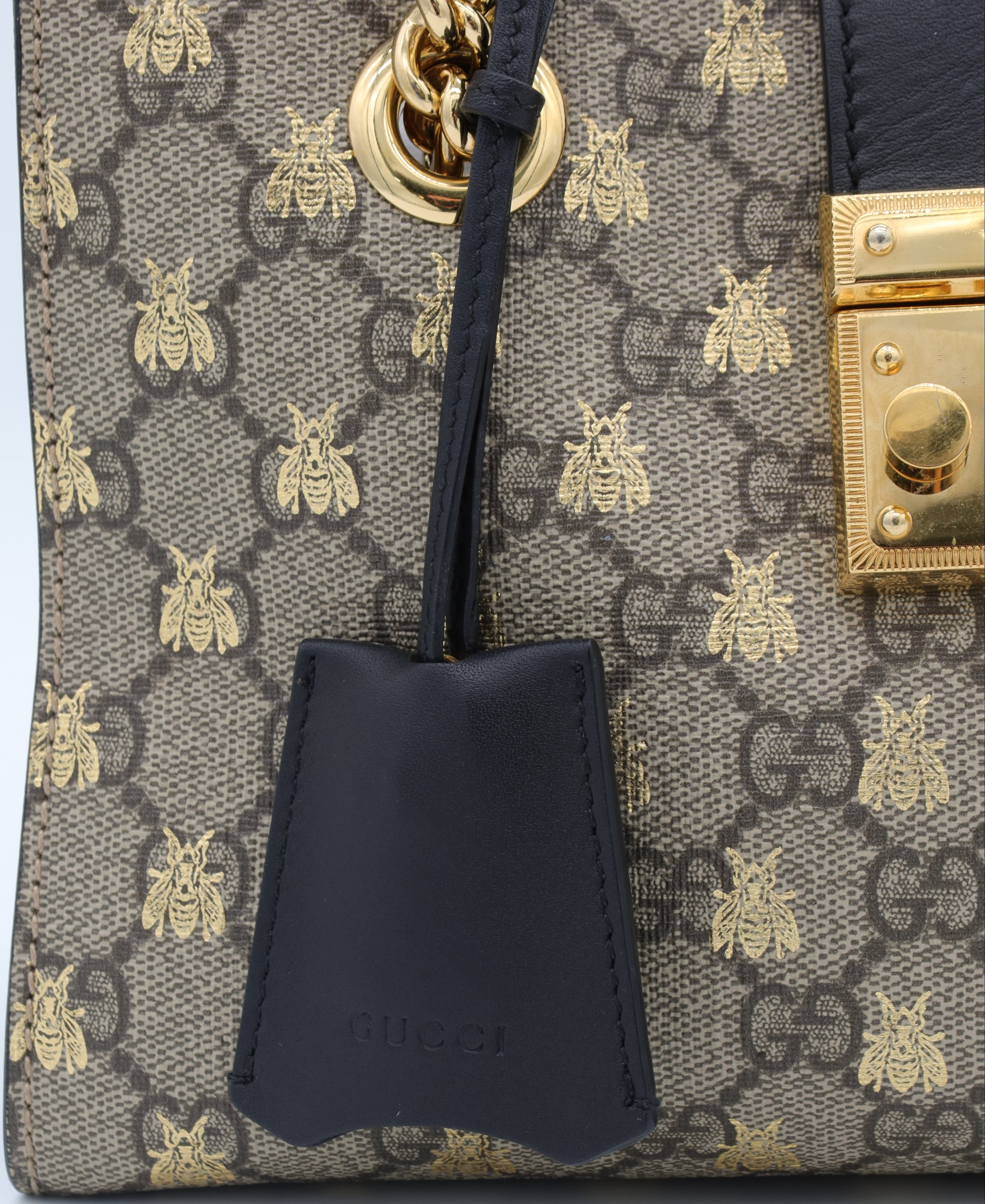 Close-up of a Gucci handbag in GG Supreme canvas with gold bee motifs, a black leather hangtag, and gold-tone hardware on a white background.