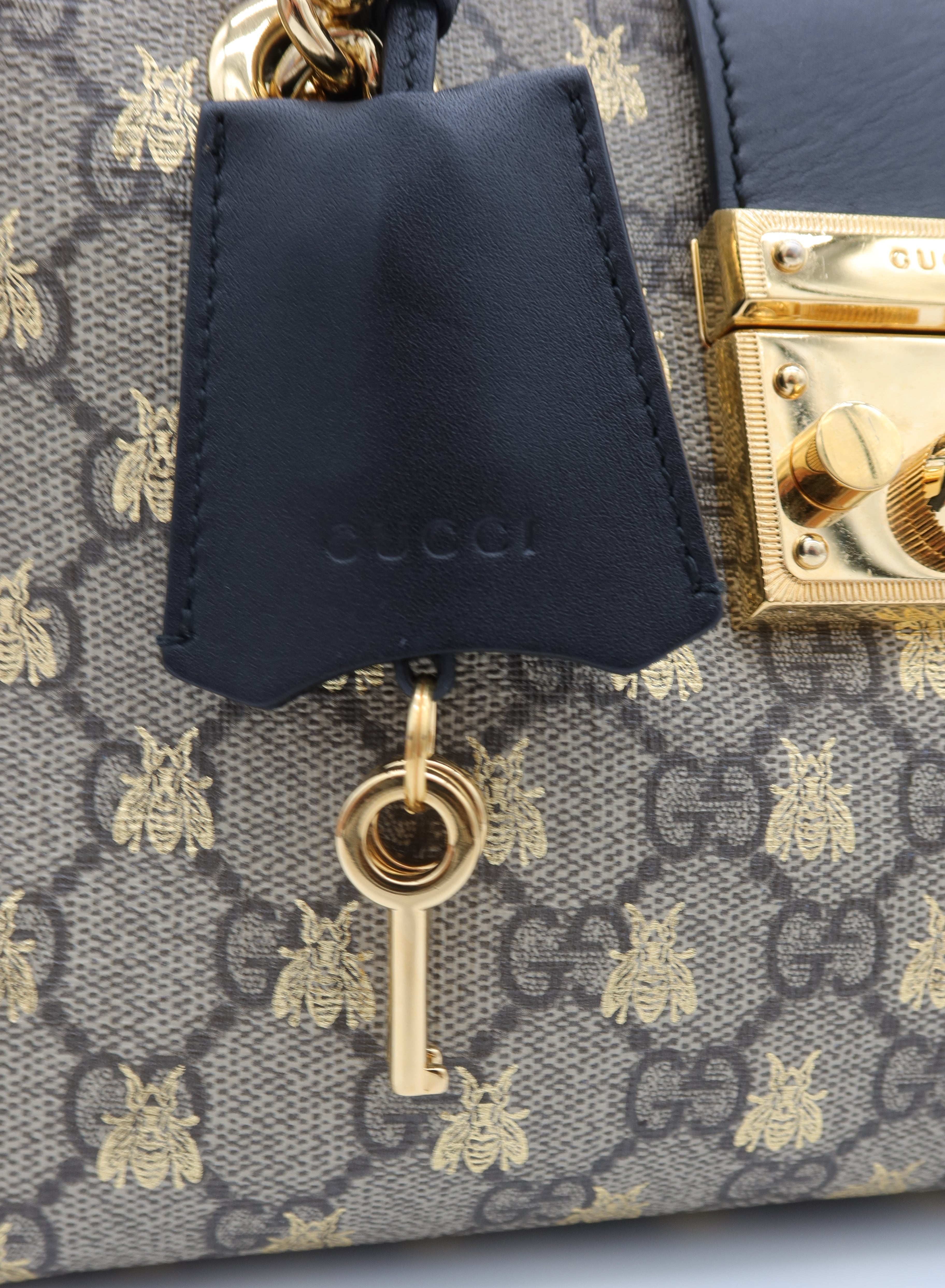 Close-up of a Gucci handbag featuring the brand's signature GG Supreme canvas with gold bee motifs, a dark navy leather hangtag, and a gold key charm.