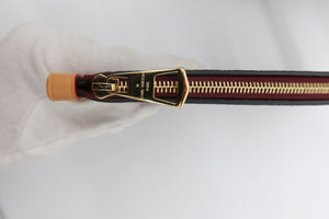 Close-up of a black leather zipper pull with gold-tone hardware and a small orange accent, held by a white gloved hand against a white background.