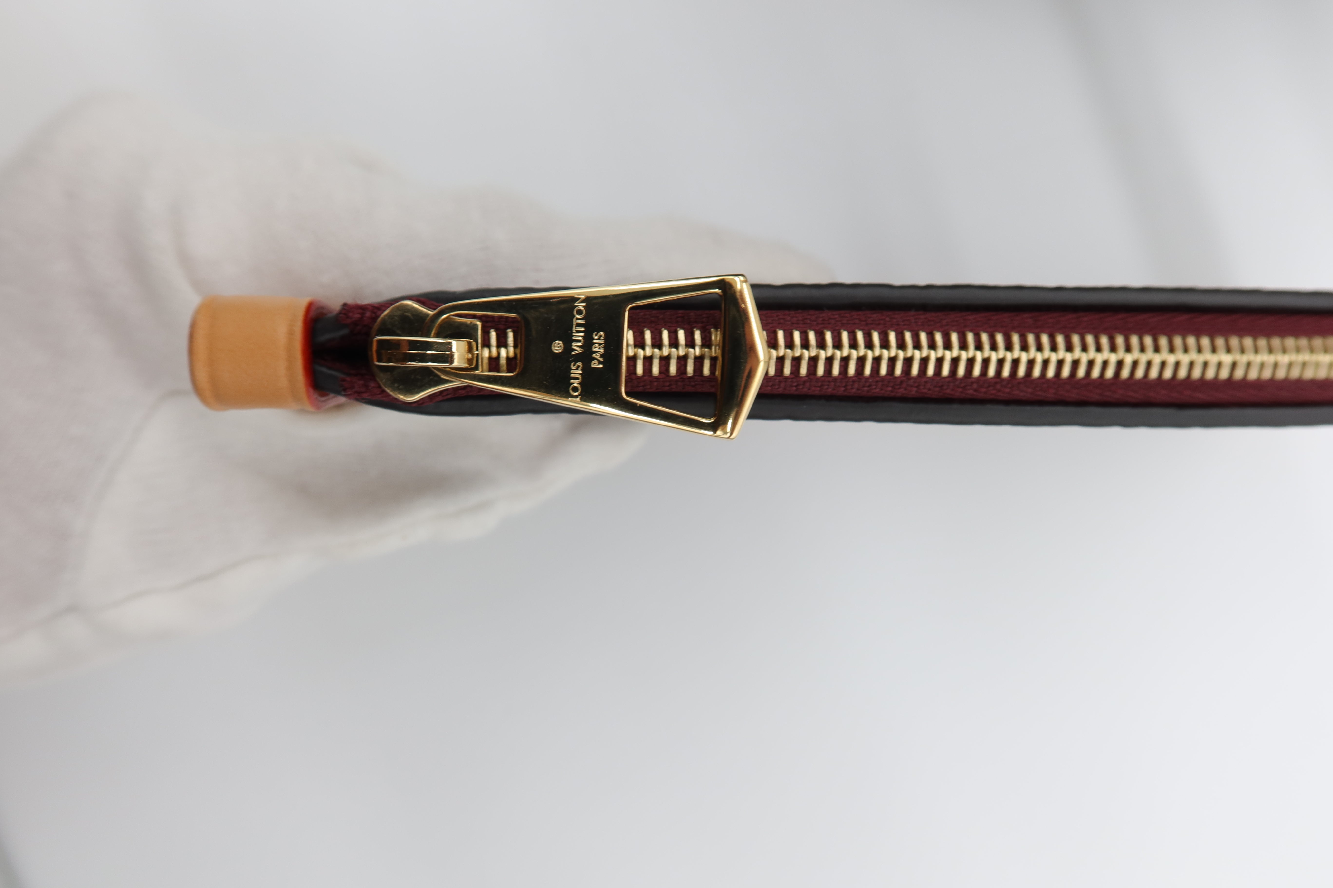 Close-up of a black leather zipper pull with gold-tone hardware and a small orange accent, held by a white gloved hand against a white background.