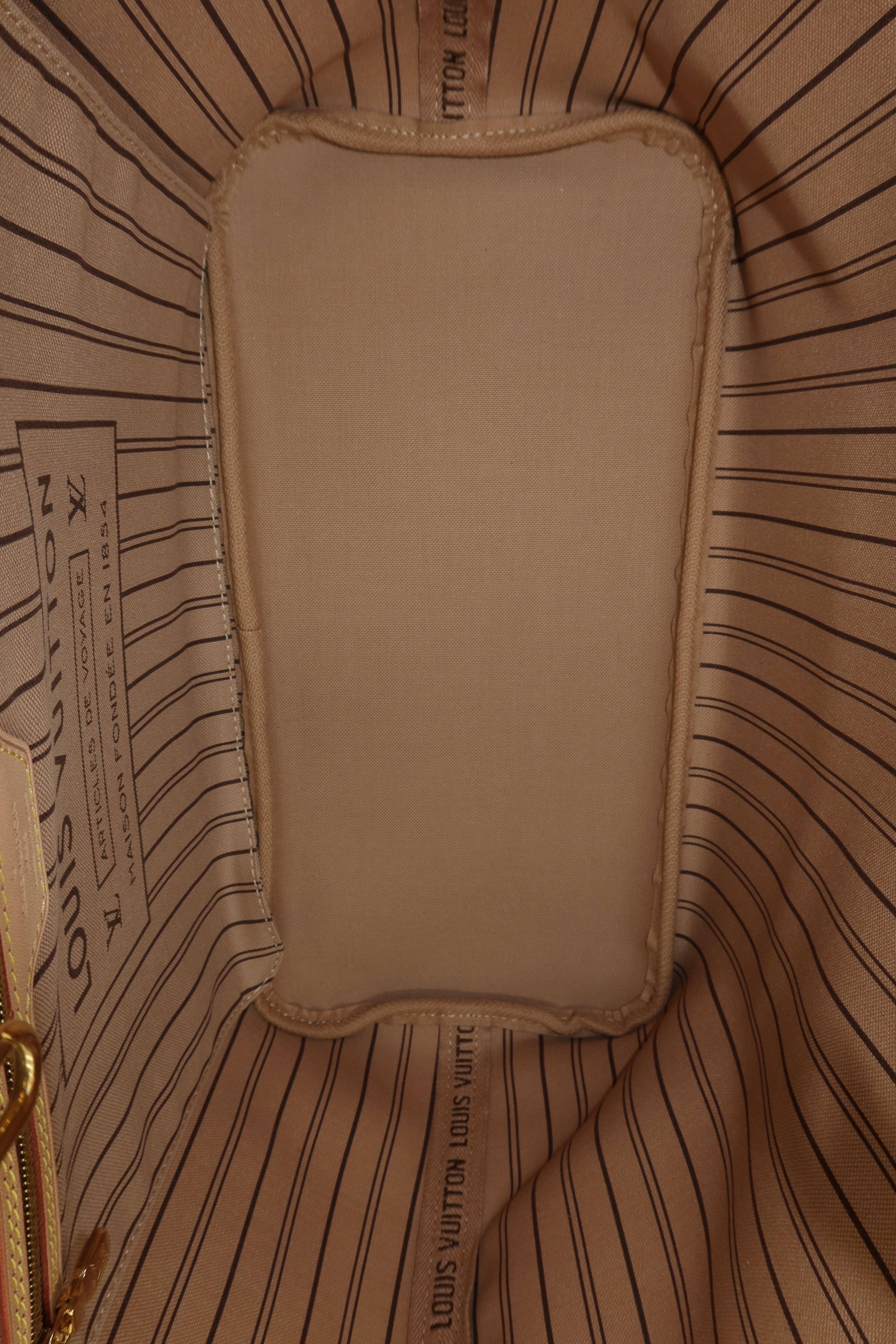 Close-up of the interior lining of a Louis Vuitton bag, showing a beige fabric insert with a rounded rectangular shape, surrounded by a beige and black striped fabric lining with Louis Vuitton branding.